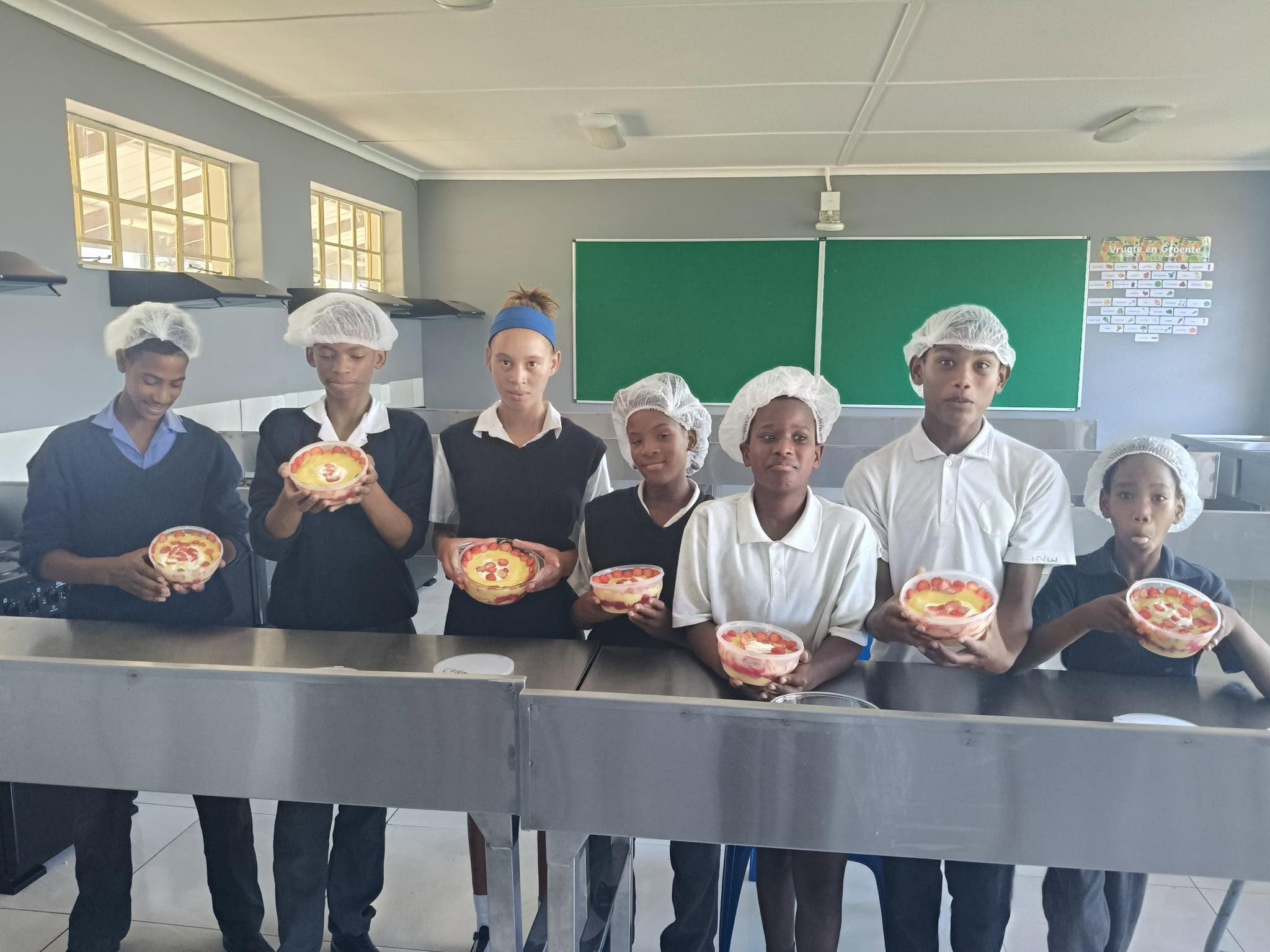 DCAPS learners in hairnets holding bowls of fruit salad they prepared in the school kitchen